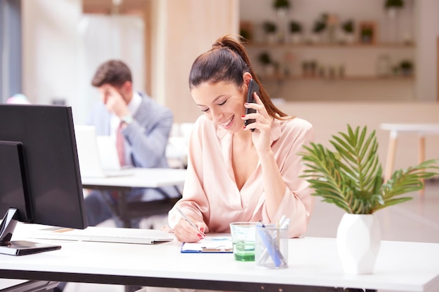 Portrait d'une femme d'affaires heureuse parlant avec quelqu'un sur son téléphone portable tout en étant assise au bureau et en train de faire de la paperasse