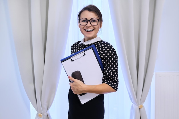 Portrait d'une femme d'affaires mature souriante et heureuse dans des verres avec des papiers, une femme regardant la caméra, à l'intérieur près de la fenêtre avec des rideaux