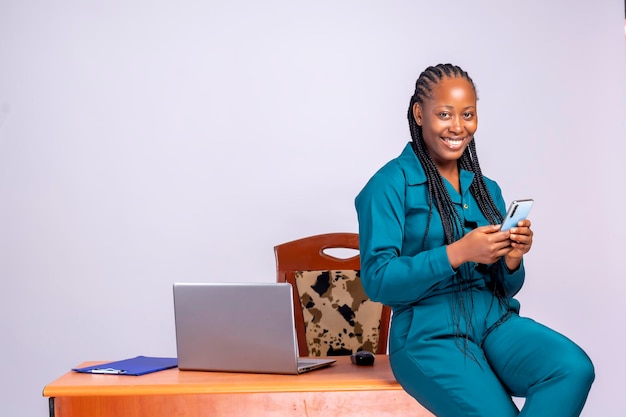 Portrait d'une femme d'affaires millénaire heureuse africaine noire utilisant son téléphone portable et son ordinateur portable, à l'intérieur, au bureau.