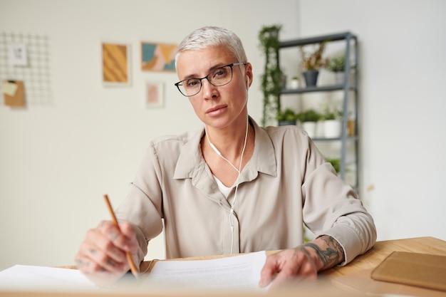 Portrait d'une femme d'affaires moderne et sérieuse d'âge moyen dans des lunettes écoutant un podcast dans des écouteurs