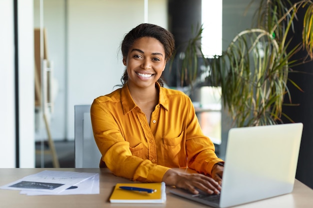 Portrait d'une femme d'affaires noire heureuse travaillant sur un ordinateur portable et souriant à la caméra assis dans l'espace de copie intérieur du bureau