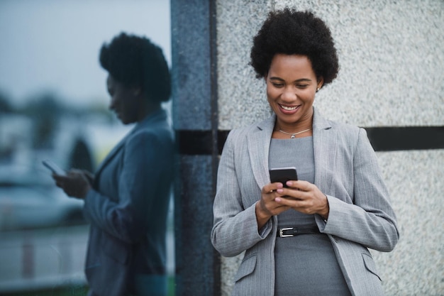 Portrait d'une femme d'affaires noire souriante utilisant l'application sur un smartphone pendant une pause rapide devant un bâtiment d'entreprise.