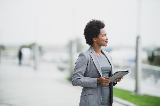 Portrait d'une femme d'affaires noire souriante utilisant une tablette numérique devant un bâtiment d'entreprise.