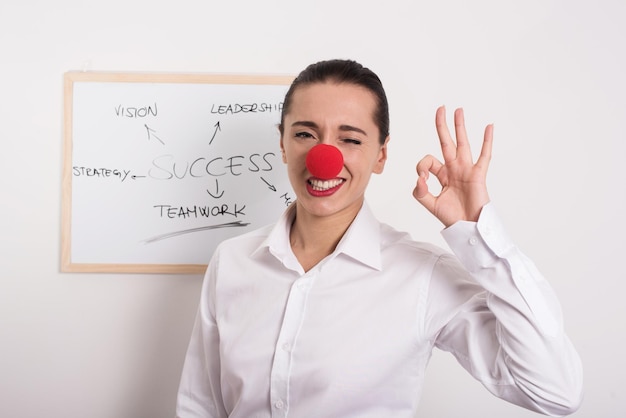 Photo portrait d'une femme d'affaires portant un nez de clown tout en faisant un geste ok au bureau