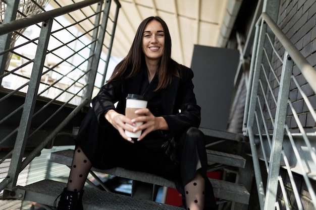 Portrait d'une femme d'affaires prospère avec une tasse de café et un large sourire assis sur les escaliers