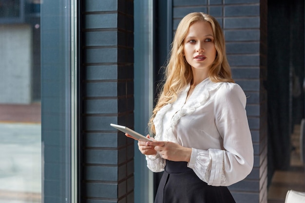 Portrait d'une femme d'affaires séduisante aux cheveux blonds en chemise classique élégante regardant de côté et tenant une tablette PC, un appareil numérique pour le travail de bureau, des applications professionnelles. debout près de la fenêtre à l'intérieur