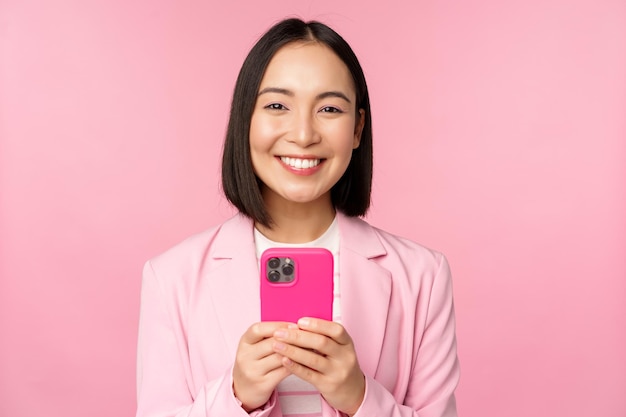 Portrait d'une femme d'affaires souriante personne morale asiatique à l'aide d'une application de téléphone mobile pour smartphone debout sur fond rose