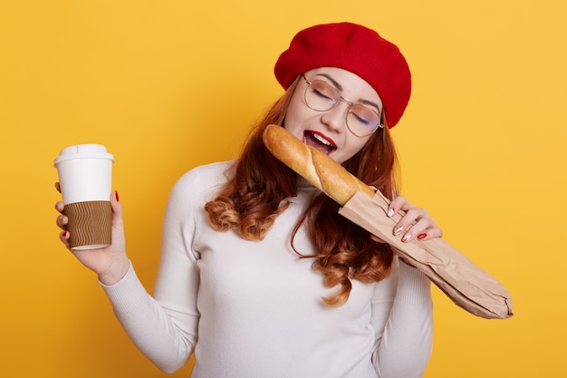 Portrait de femme affamée heureuse tenant un sac en papier avec une baguette de pain blanc et mordant sur jaune