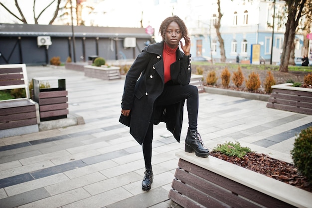 Portrait d'une femme africaine aux cheveux bouclés portant un manteau noir à la mode et un col roulé rouge posant en plein air