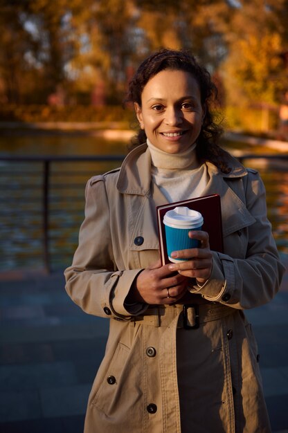 Portrait d'une femme africaine joyeuse avec une tasse de café à emporter et un livre à main souriant regardant la caméra sur le fond du lac du parc en automne avec des feuilles qui tombent. Loisirs de plein air automnaux. des loisirs