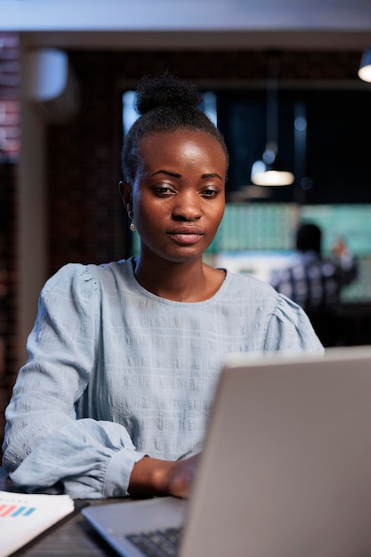 Portrait d'une femme afro-américaine confiante assise au bureau tout en utilisant un ordinateur portable pour effectuer une analyse de marché en temps réel. Négociant en actions Forex dans l'espace de travail de l'agence examinant les statistiques d'investissement.