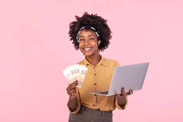 Portrait d'une femme afro-américaine excitée tenant des billets d'argent avec un ordinateur portable isolé sur un fond rose Portraitd'une jeune femme joyeuse tenant des billits d'argent et un ordinateur laptop