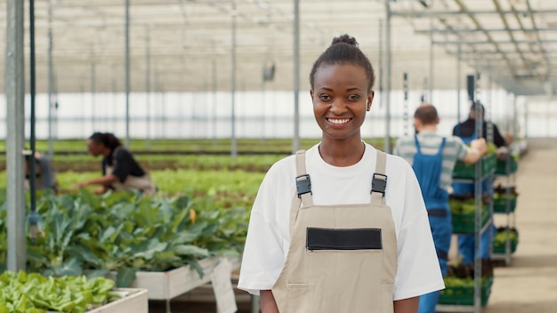 Portrait d'une femme afro-américaine posant heureuse dans des cultures biologiques et une ferme de légumes tandis que divers travailleurs poussent des caisses avec des cultures. Ouvrier agricole souriant dans une serre debout dans un environnement hydroponique.