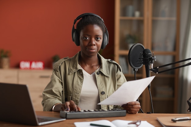 Portrait de femme afro-américaine souriante regardant la caméra et portant des écouteurs tout en composant de la musique en studio d'enregistrement, copiez l'espace