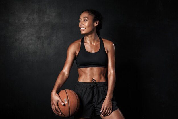 Portrait d'une femme afro-américaine souriante en tenue de sport tenant un basket-ball isolé sur fond noir