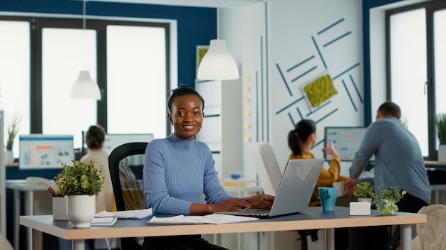 Portrait d'une femme afro-américaine souriante travaillant sur la marge bénéficiaire et les statistiques de vente à l'aide d'un ordinateur portable assis au bureau dans un bureau de démarrage occupé. Employé d'entreprise occasionnel satisfait du résultat de son travail.