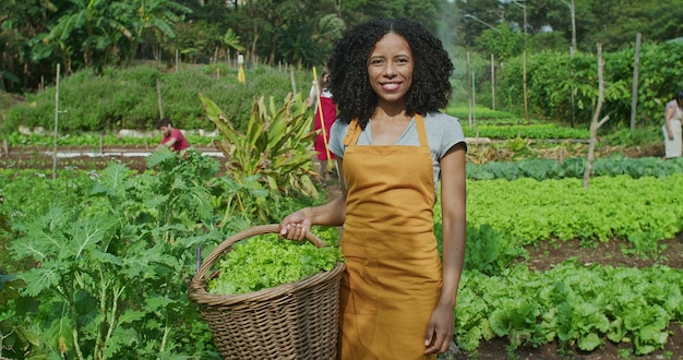 Photo portrait d'une femme afro-américaine tenant un panier de laitues biologiques debout dans une ferme communautaire regardant la caméra groupe de personnes en arrière-plan travaillant dans un jardin communautaire cultivant de la nourriture