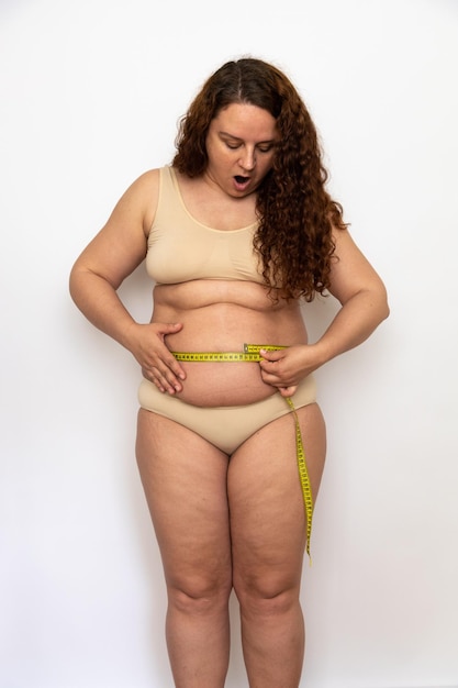 Portrait d'une femme d'âge moyen dodue choquée aux cheveux bouclés portant des sous-vêtements beiges mesurant la taille avec du ruban jaune