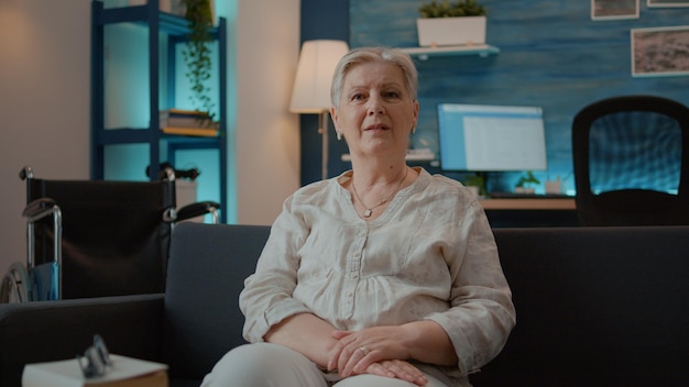 Portrait d'une femme âgée avec une blessure physique assise sur un canapé. Personne regardant la caméra et assise à côté d'un fauteuil roulant dans le salon, utilisant un équipement de mobilité pour le transport.