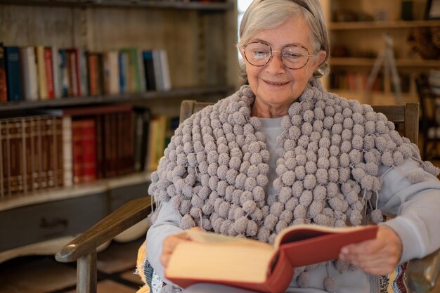 Portrait d'une femme âgée dans des verres assis sur un fauteuil lisant un livre papier roman best-seller