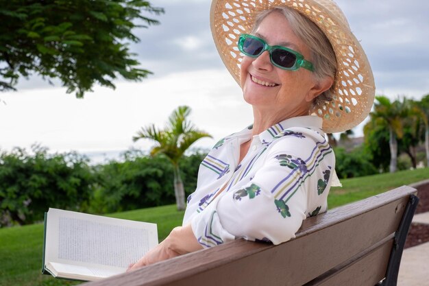 Photo portrait d'une femme âgée gaie avec des lunettes de soleil vertes et un chapeau assis sur un banc dans un parc public lisant un livre caucasien dame regardant la caméra en souriant