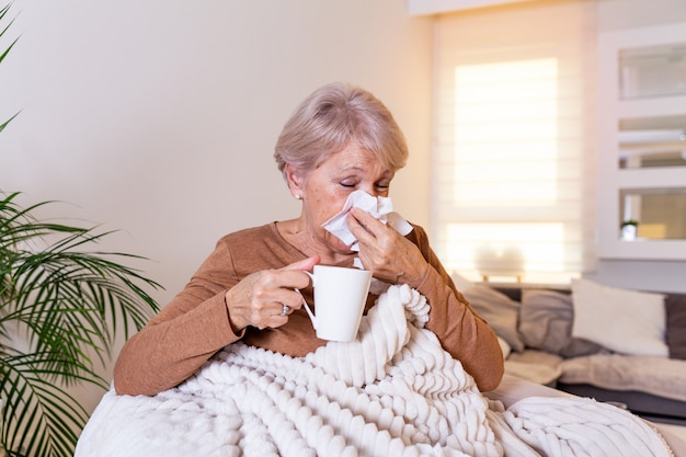 Portrait de femme âgée malade pris froid, se sentir malade et éternuer dans une lingette en papier recouvert d'une couverture en essuyant le nez.