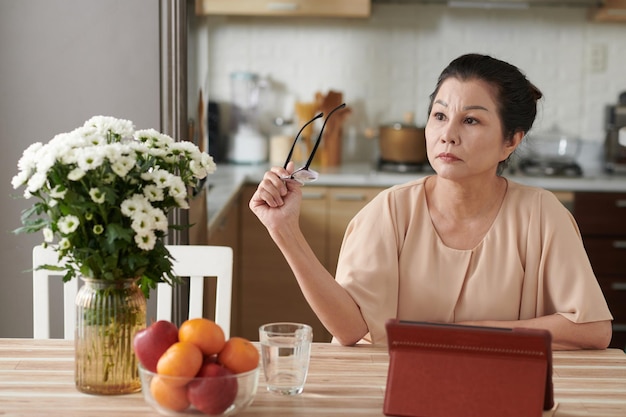 Portrait d'une femme âgée pensive assise à la table de la cuisine avec une tablette numérique et enlevant des lunettes