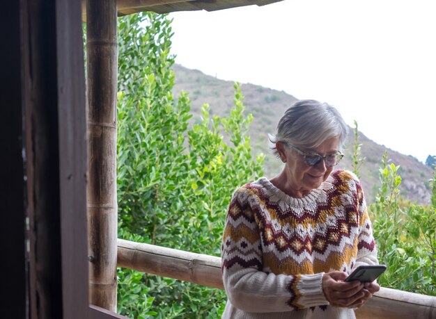 Portrait d'une femme âgée en plein air sur un balcon portant un pull d'hiver et buvant à l'aide d'un téléphone portable. Femme âgée détendue aux cheveux gris profitant de la nature et du chalet de montagne