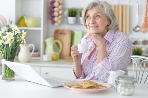 Portrait d'une femme âgée prenant son petit déjeuner avec un ordinateur portable