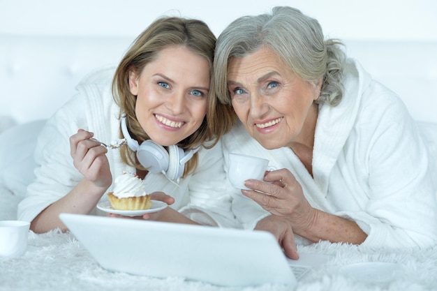 Portrait d'une femme âgée avec sa fille adulte mangeant un délicieux gâteau, buvant du thé et regardant quelque chose sur un ordinateur portable