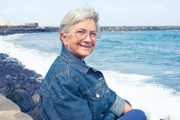 Portrait d'une femme âgée souriante aux cheveux blancs assise sur la plage portant des lunettes et une veste en jean Jolie femme de 70 ans profitant du bon temps et des vacances