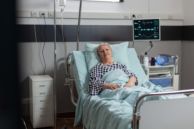 Portrait d'une femme âgée souriante regardant la caméra dans un lit d'hôpital pariant un traitement par le biais d'un sac d'égouttement iv. Respirer avec l'aide d'un masque à oxygène pendant la récupération de la maladie.