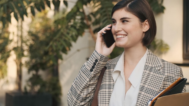 Portrait d'une femme agent immobilier parlant au téléphone et souriant en attendant les clients dans la rue de la ville Expression du visage heureux