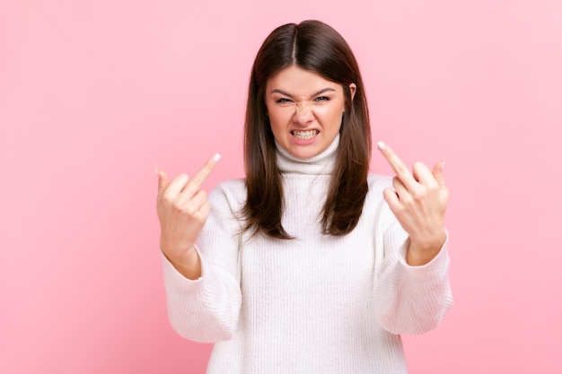 Portrait d'une femme agressive en colère debout et montre un signe de baise avec les deux mains dans l'appareil photo, portant un pull blanc de style décontracté. Tourné en studio intérieur isolé sur fond rose.