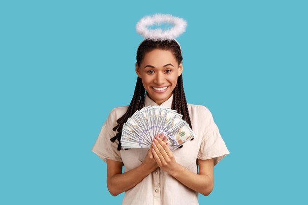 Portrait d'une femme angélique souriante et satisfaite avec des dreadlocks et des plumes sur la tête tenant un éventail de billets en dollars portant une chemise blanche Prise de vue en studio intérieure isolée sur fond bleu