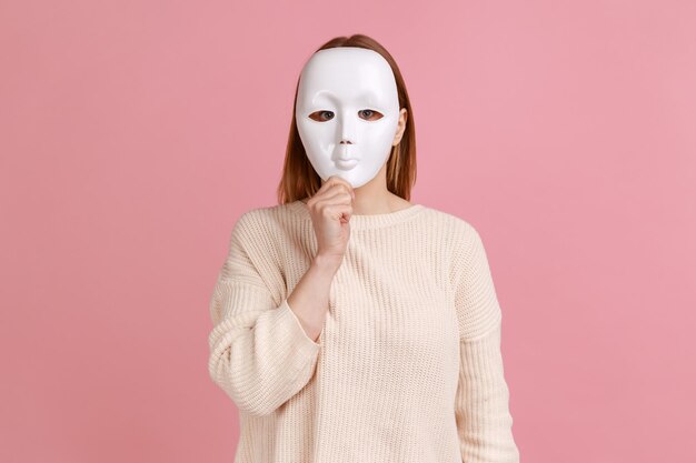 Portrait d'une femme anonyme inconnue couvrant son visage avec un masque blanc cachant sa vraie personnalité anonymat portant un chandail blanc Prise de vue en studio intérieure isolée sur fond rose