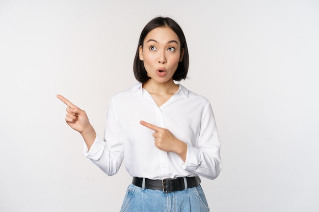 Portrait d'une femme asiatique adulte séduisante pointant vers la gauche avec un sourire heureux montrant une bannière ou un logo de côté debout sur fond de studio blanc