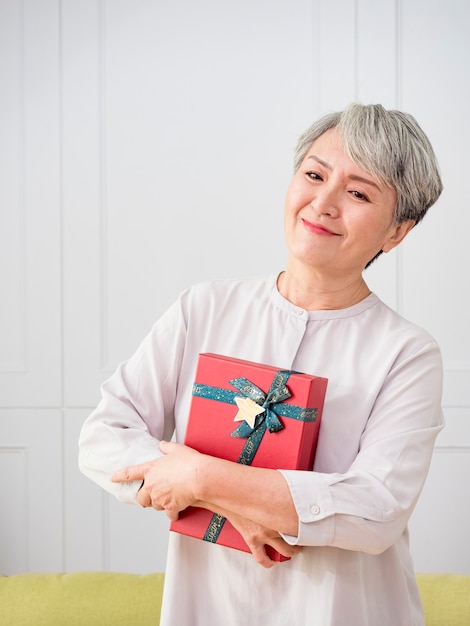 portrait d'une femme asiatique âgée tenant une boîte-cadeau, souriante à la maison.