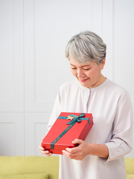 portrait d'une femme asiatique âgée tenant une boîte-cadeau, souriante et regardant vers le bas à la maison.