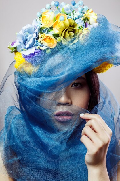 Portrait de femme asiatique avec chapeau floral et voile bleu regardant la caméra sur fond gris clair. tourné en studio intérieur.