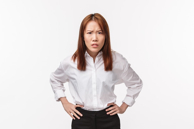 Portrait de femme asiatique déçue et stricte tenir la main sur la taille et se pencher en avant, froncer les sourcils avec blâme, accuser la personne d'être impoli, réprimander l'employé désobéissant sur le mur blanc