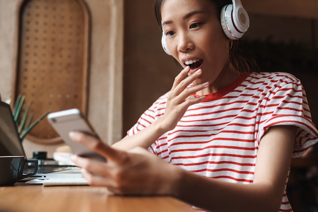 Portrait d'une femme asiatique excitée écoutant de la musique avec des écouteurs et tenant un téléphone portable dans un café à l'intérieur