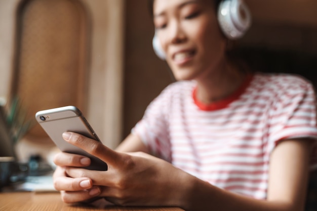 Portrait d'une femme asiatique heureuse écoutant de la musique avec des écouteurs et tenant un téléphone portable dans un café à l'intérieur