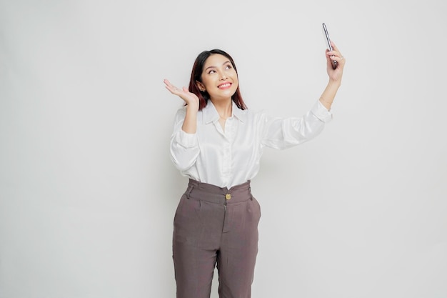 Un portrait d'une femme asiatique heureuse portant une chemise blanche et tenant son téléphone isolé par fond blanc