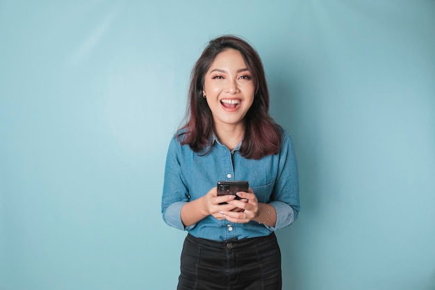 Un portrait d'une femme asiatique heureuse sourit et tient son smartphone portant une chemise bleue isolée par un fond bleu