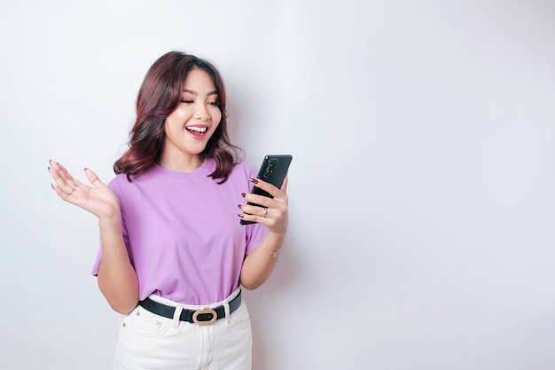 Un portrait d'une femme asiatique heureuse sourit et tient son smartphone portant un t-shirt violet lilas isolé par un fond blanc