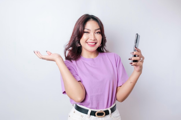 Un portrait d'une femme asiatique heureuse sourit et tient son smartphone portant un t-shirt violet lilas isolé par un fond blanc