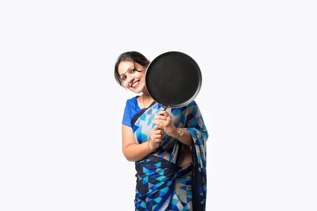 Portrait d'une femme asiatique indienne en sari dans la cuisine, tenant une louche en bois, un belan, une spatule ou une casserole, isolée contre un mur blanc