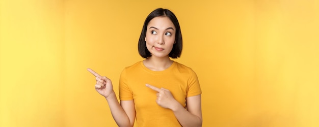 Portrait d'une femme asiatique intriguée regardant et pointant du doigt à gauche une publicité montrant quelque chose d'intéressant debout sur fond jaune