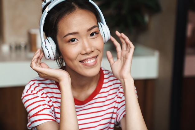 Portrait d'une femme asiatique joyeuse écoutant de la musique avec des écouteurs alors qu'elle était assise dans un café à l'intérieur